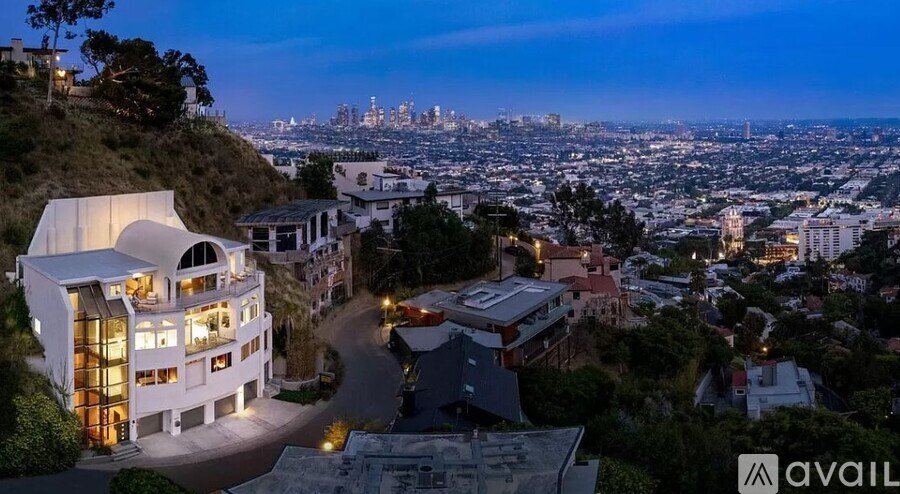 A modern house with a curved roof is lit up at night with a cityscape in the background.
