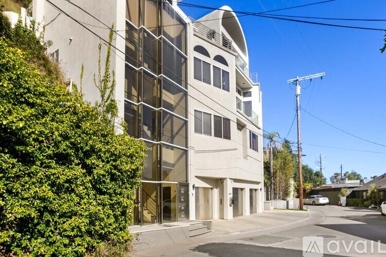 A modern building with a curved corner and a glass facade is surrounded by greenery and a clear sky.
