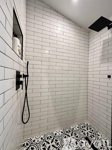 A white tiled shower with a black shower head and a black and white patterned shower mat.