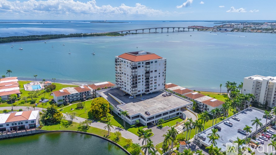 A large white building with a red roof is surrounded by a parking lot and palm trees.