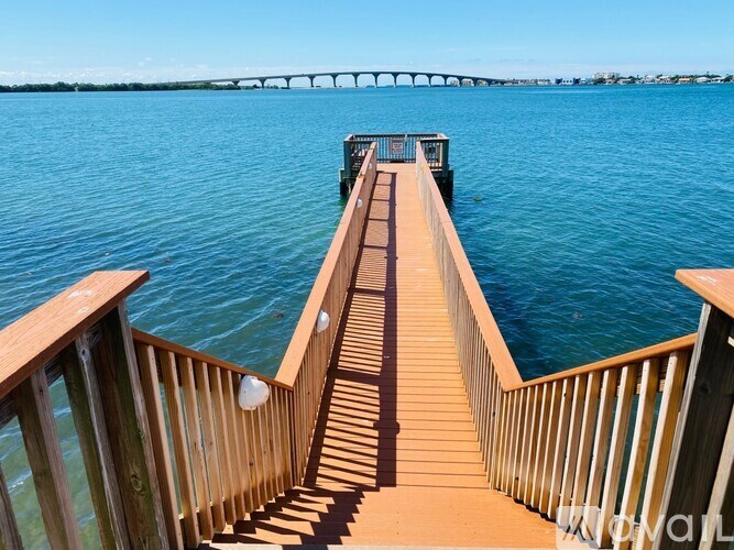 A wooden pier extends into a blue body of water with a bridge in the distance.