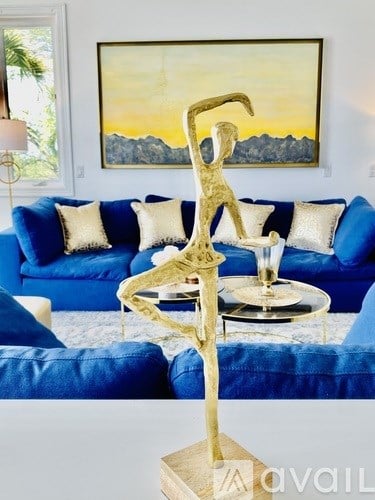 A gold sculpture of a person holding a glass is in the foreground of a living room with a blue couch and a painting on the wall.