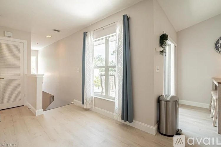 A room with a large window and a curtain, a trash bin, and a wall clock.