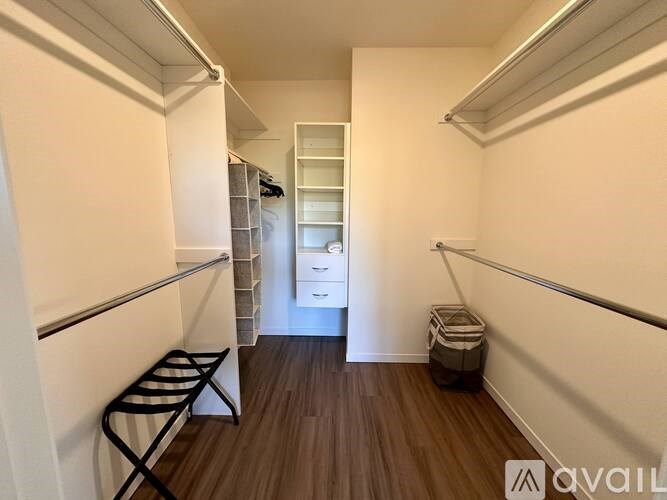 A room with white walls and a wooden floor, featuring a white wardrobe, a black chair, a white dresser, and a brown basket.