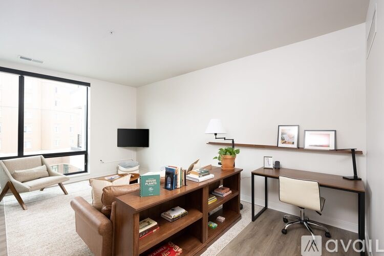 A living room with a brown leather couch, a wooden bookshelf, a white chair, and a flat-screen TV mounted on the wall.