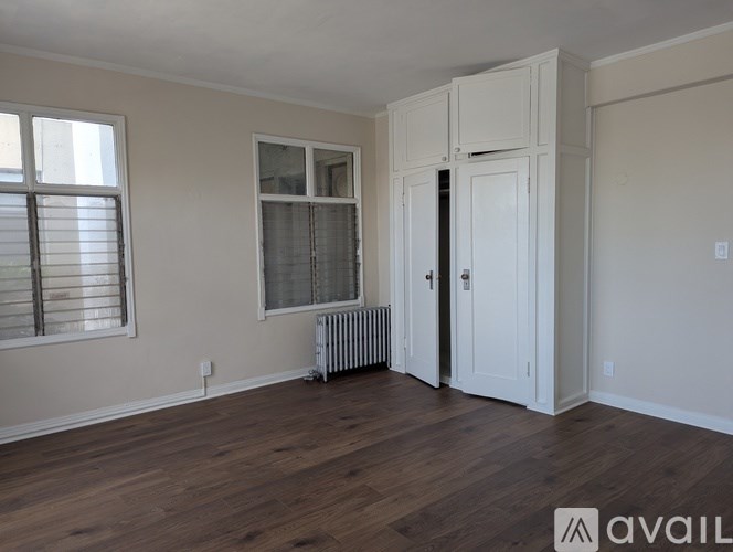 A room with wooden floors and white walls, featuring a window with blinds, a radiator, and a door leading to another room.