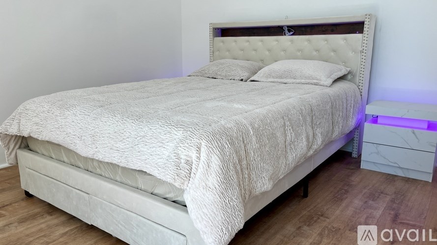 A bed with a white comforter and two pillows sits in a room with a wooden floor and a white wall.