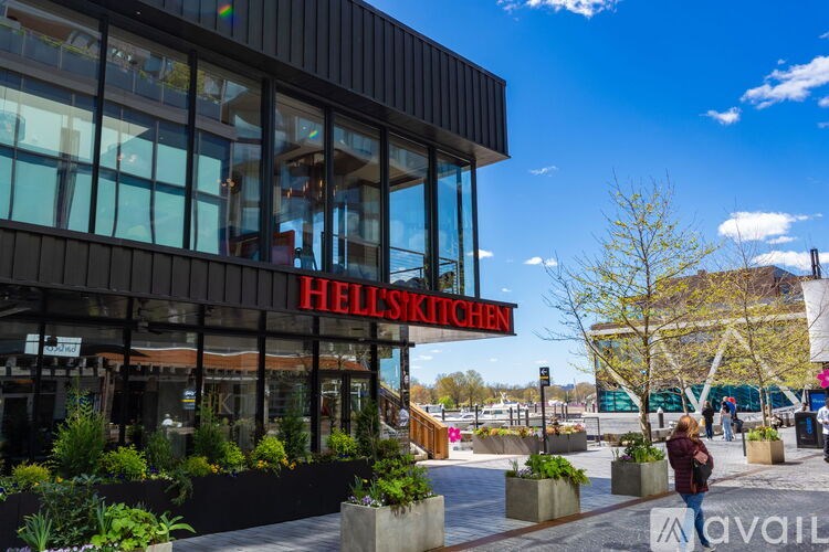 A modern building with a red sign that says "HELL'S KITCHEN" is surrounded by plants and people are walking by.