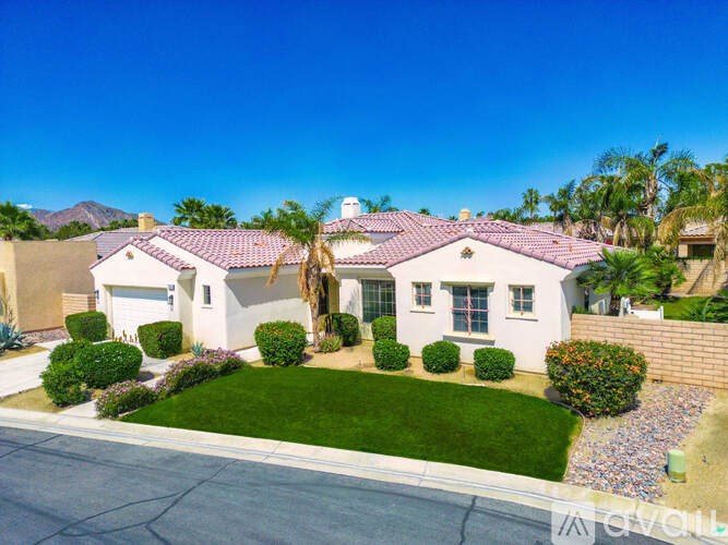 A white house with a red tile roof is surrounded by a well-manicured lawn and palm trees.
