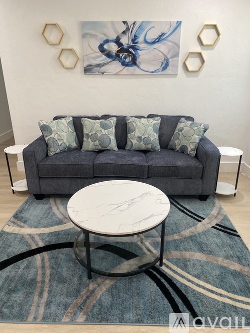 A grey couch with four pillows and a white coffee table in front of a painting.