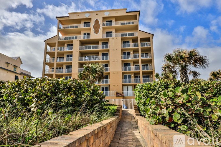 A tall beige building with balconies and a brick pathway leading to the entrance.