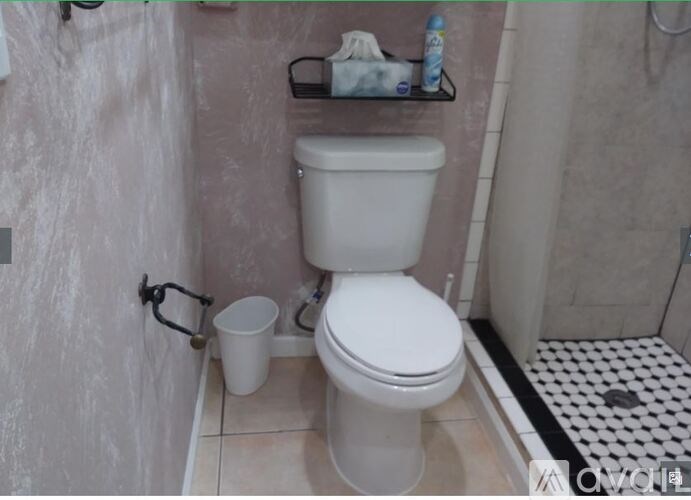 A white toilet in a bathroom with a white bucket and a roll of toilet paper on the shelf.