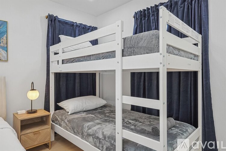 A white bunk bed with a grey blanket and a white nightstand with a lamp on it.