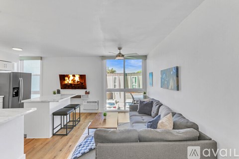 A modern living room with a grey sofa and a kitchen area with a fridge and bar stools.