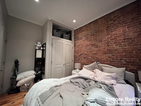 A bedroom with a brick wall and a bed with a grey comforter.