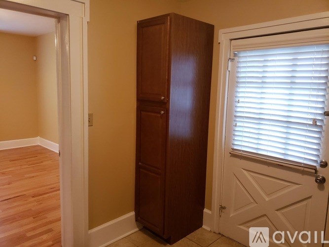 A tall brown wooden wardrobe stands in a room with a white door and window with blinds.