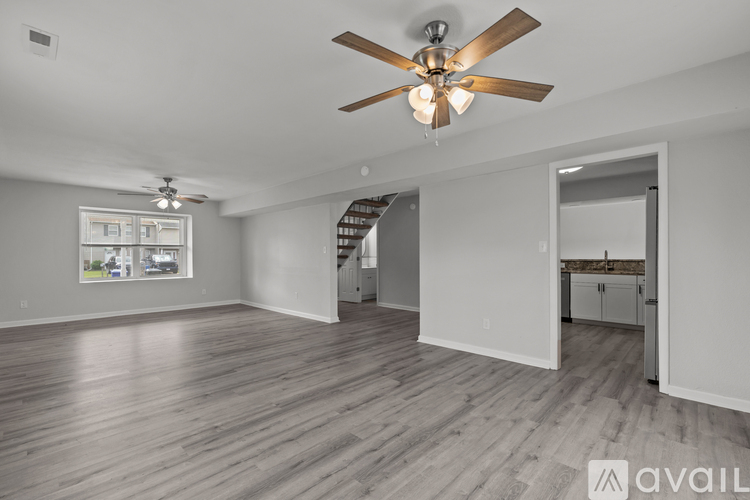 A spacious room with a ceiling fan and light fixture, hardwood flooring, and a view of a kitchen area through an open door.