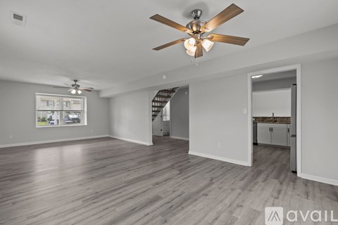 A spacious room with a ceiling fan and light fixture, hardwood flooring, and a view of a kitchen area through an open door.