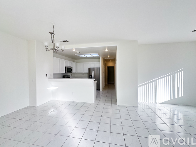 A white tiled room with a chandelier and a kitchen area in the background.