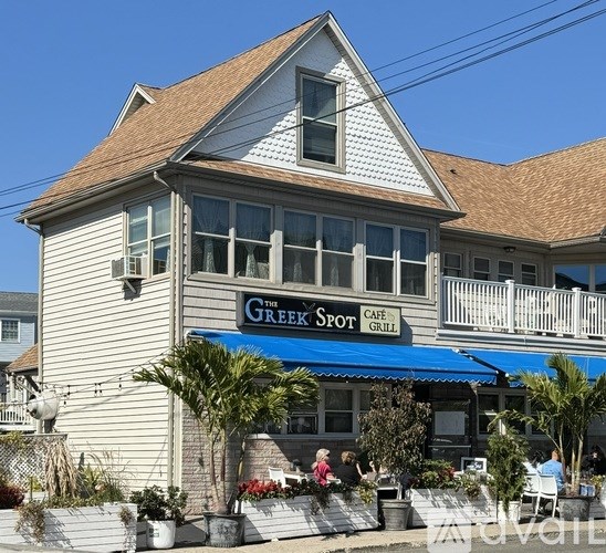 A cafe named The Greek Spot has a blue awning and is surrounded by potted plants.