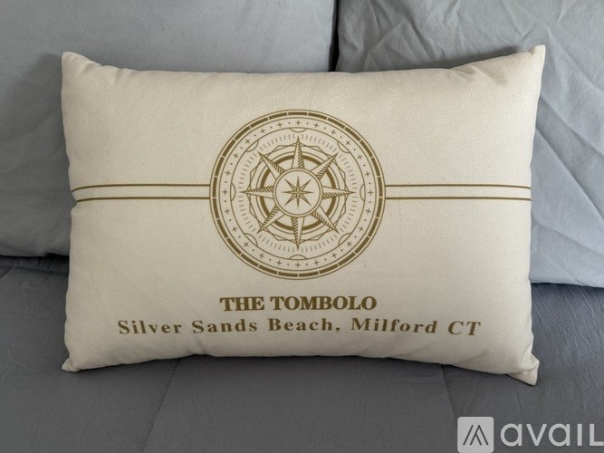 A decorative pillow with a compass design and the text "THE TOMBOLO Silver Sands Beach, Milford CT" is displayed on a bed.