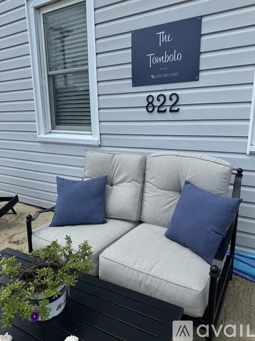 A white couch with two blue pillows sits on a black table outside a house with a sign that says "The Tombolo" and the number 822.