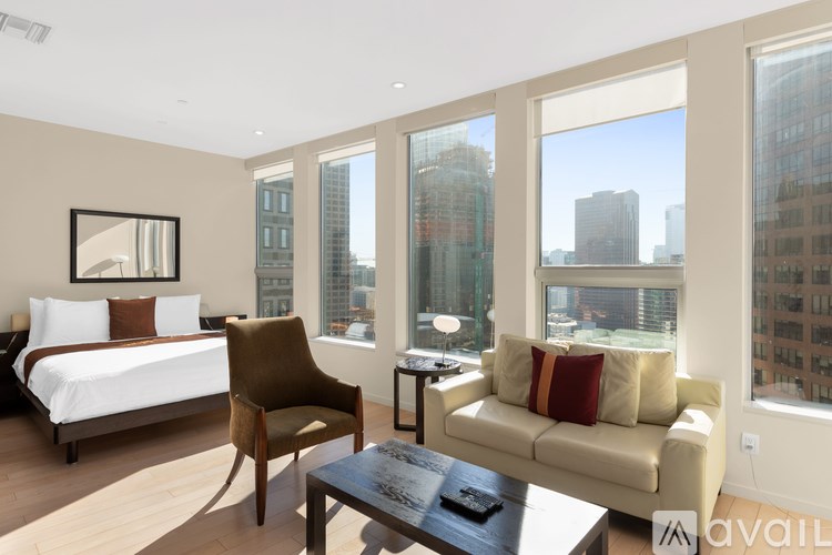 A bedroom with a bed, chair, and couch with a city view through the windows.