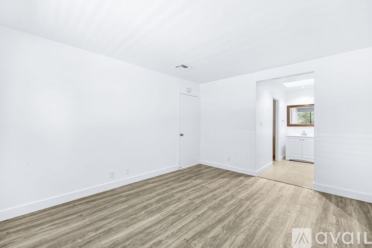 A room with wooden flooring and white walls, with a door on the left and a hallway on the right.