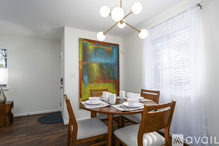 A dining room with a wooden table and chairs, a painting on the wall, and a window with white curtains.