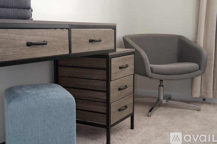 A grey chair sits next to a wooden desk with drawers and a blue ottoman.