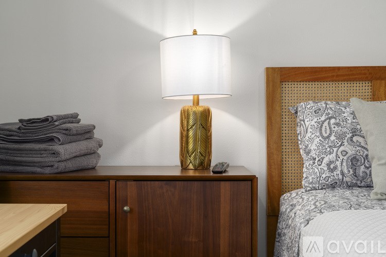 A lamp with a gold base and a white lampshade is on a wooden cabinet.