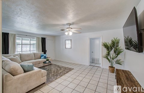 A living room with a beige couch, a rug, a ceiling fan, and a flat-screen TV mounted on the wall.