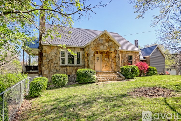 A house with a stone facade and a brown roof is surrounded by greenery.