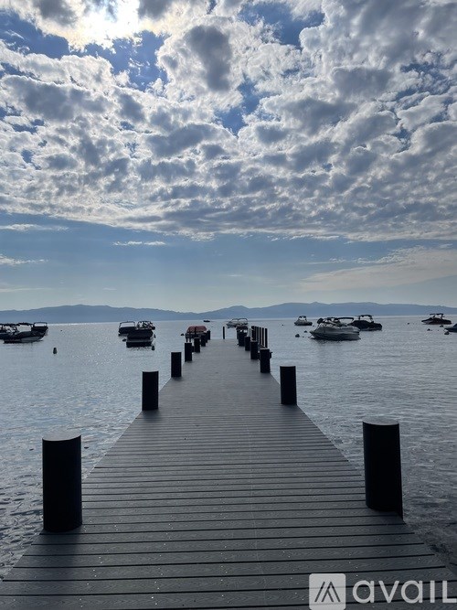 A wooden pier extends into a calm body of water with boats and mountains in the distance under a cloudy sky.
