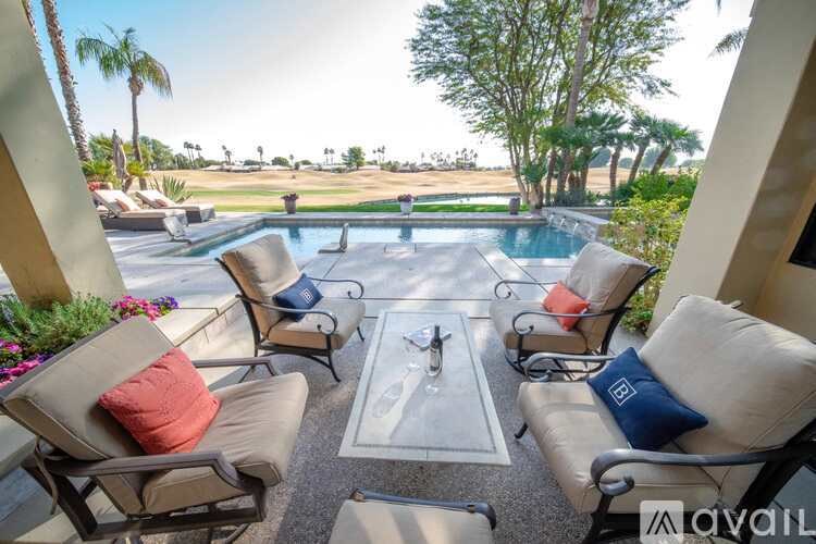 A patio with chairs and a table is surrounded by a pool and palm trees.