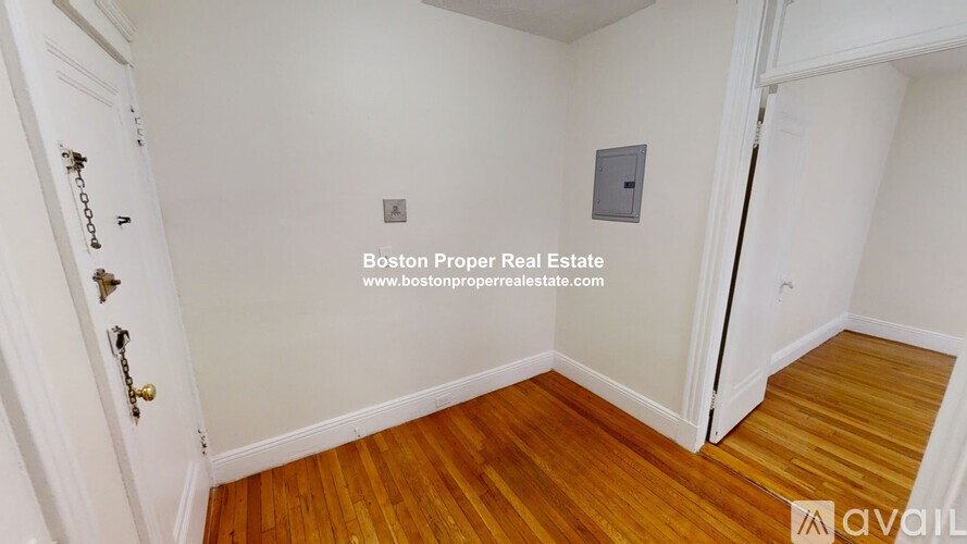 A room with wooden flooring and white walls, with a door on the left and a small electrical box on the wall.