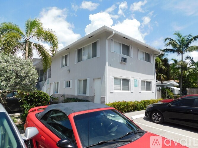 A red car is parked in front of a white building with palm trees in the background.