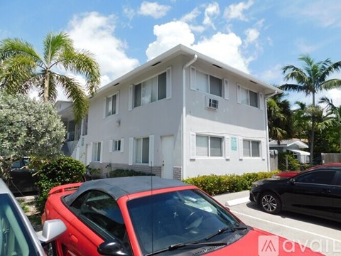 A red car is parked in front of a white building with palm trees in the background.