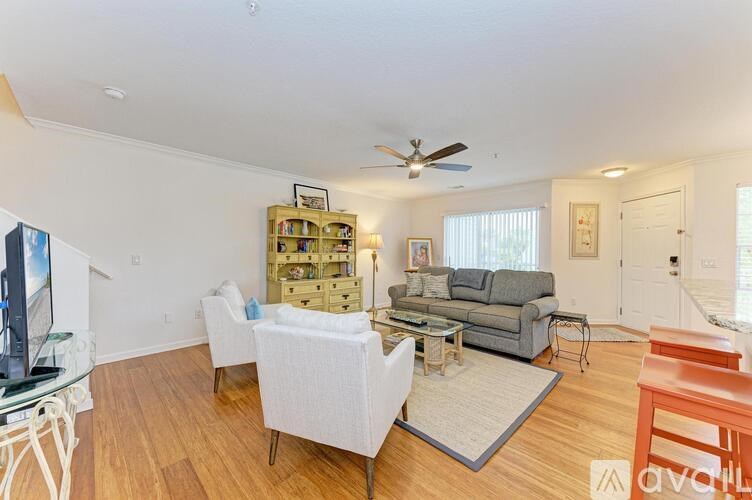 A living room with a grey couch, a white chair, a white coffee table, a white rug, a white TV stand, a white bookshelf, a white ceiling fan, and a white door.