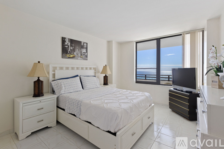 A bedroom with a large bed, a nightstand with a lamp, and a window overlooking the ocean.
