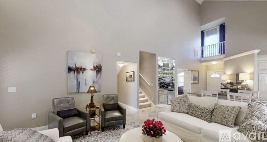 A living room with a grey couch, a grey and white rug, a grey and black chair, a table with a vase of red flowers, a painting on the wall, and a staircase leading to the upper floor.