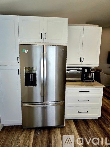 A stainless steel refrigerator with a water and ice dispenser in the middle of the door stands in a kitchen with white cabinets and a wooden floor.