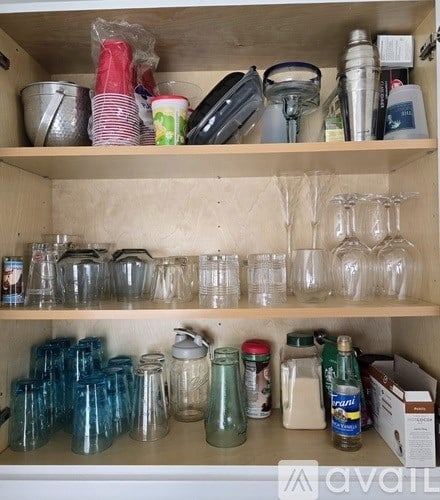 A kitchen cabinet is full of glasses, jars, and other items.