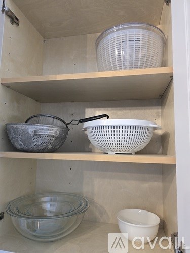 A kitchen cabinet with three shelves containing a colander, a glass bowl, and a white bowl.