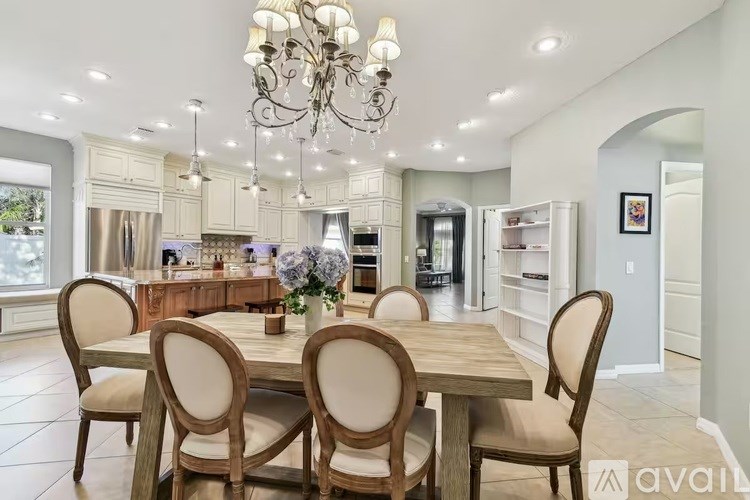A dining room with a wooden table and chairs, a chandelier, and a kitchen in the background.