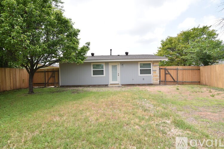 A small house with a grey roof and a grey door is surrounded by a fence and a tree.