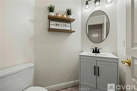 A bathroom with a white toilet, a grey cabinet, a round mirror, and a shelf with plants and candles.