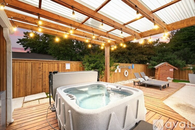A hot tub is in the middle of a wooden deck under a roof with lights strung above it.