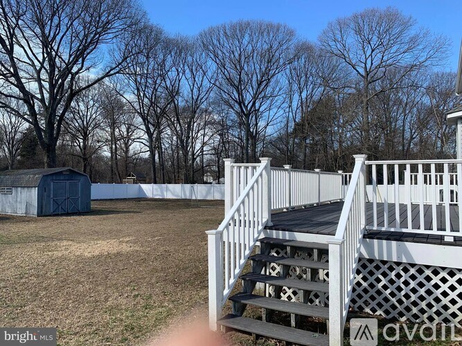 A white deck with a lattice railing is in the foreground of a grassy field with a shed and trees in the background.