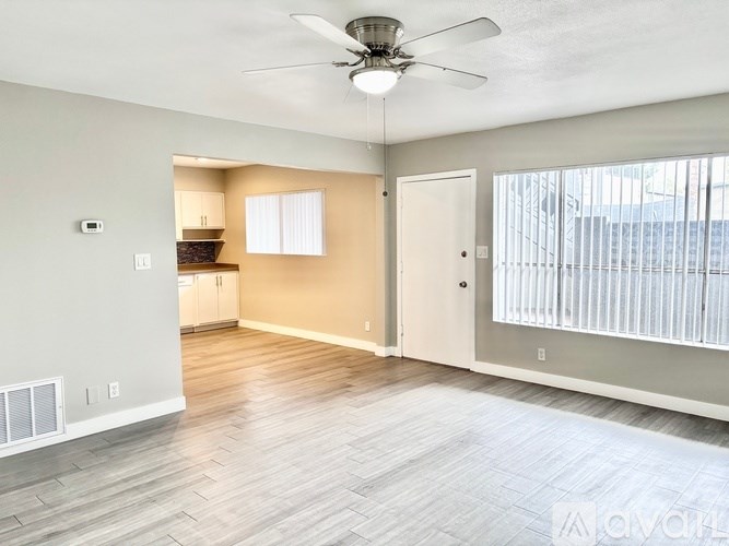 A spacious room with a ceiling fan and light fixture, hardwood flooring, and a view of a parking lot through a window.